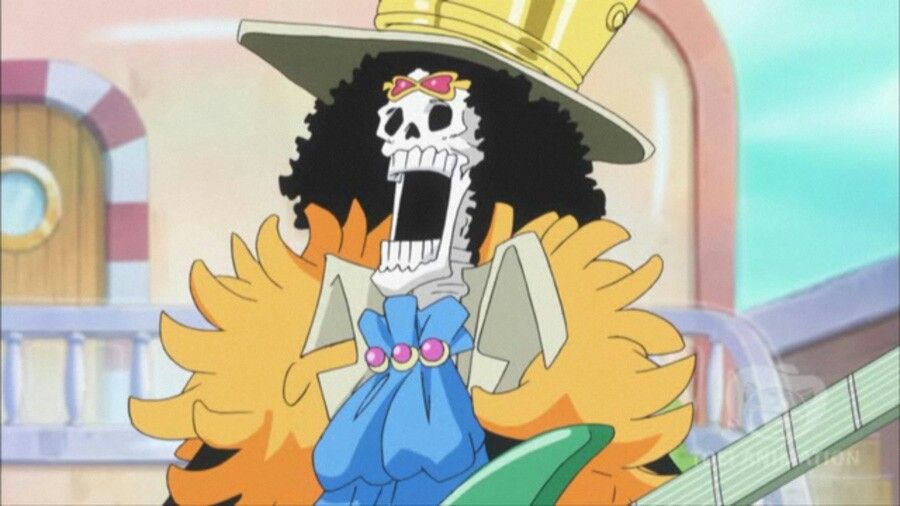 Brook, tem cabelos pretos encaracolados, usando um chapéu branco e dourado, um cachecol laranja felpudo, uma blusa branca e uma espécie de gravata azul